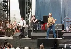 U2 playing on an outdoor stage. The Edge is on the left playing guitar, Bono in the centre with a microphone, and Adam Clayton on the right playing bass guitar. A drum set is partially visible on the right side.