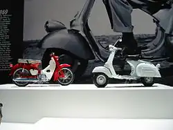 A 1960s Italian scooter and a 1960s single cylinder Japanese motorcycle in front of a giant image on the wall of the front wheel and fender of a 1960s Italian scooter.