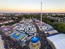 A panoramic view of the Schweinfurt Volksfest festival grounds in 2025, photographed from the Ferris wheel. The photo shows the festival in the evening with rows of booths, food vendors and rides spread across the grounds. A brightly lit drop tower ride stands near the centre-right. The area is surrounded by green trees and urban buildings, highlighting the festival central location within the city of Schweinfurt.