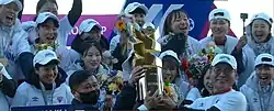 A group of female football players, along with their managers, lift a large gold trophy, smiling joyfully.
