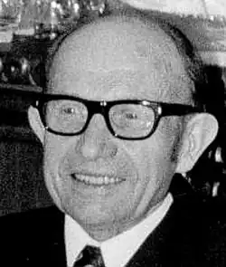 Black and white Press shot of a smiling, elderly, balding, clean-shaven and bespectacled man, with prominent ears
