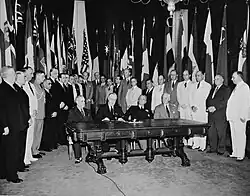 Franklin D. Roosevelt and three other men seated at a table, surrounded by many other men and flags
