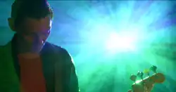 A screenshot from the music video; Richard Jones playing Bass, behind him is as an image projector creating a lens flare.