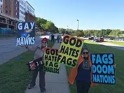 Two women standing on a university campus sidewalk, holding placards reading "Gay Hawks", "God Hates Fag Enablers", "God Hates Fags", and "Fags Doom Nations"