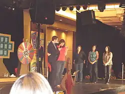 Marty Lublin interviewing a woman on a small stage. Three other women and a roulette-style wheel are also visible on the stage.