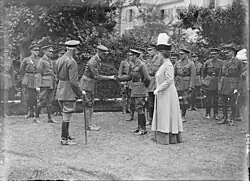 A black-and-white photograph showing a uniformed man on the left who is stooping slightly forward as he holds the hand of a man on the right, who is the king. Also in the photograph are many other uniformed men, forming a group mostly behind the two main subjects. A woman wearing a feathered hat stands to the left side of the king.